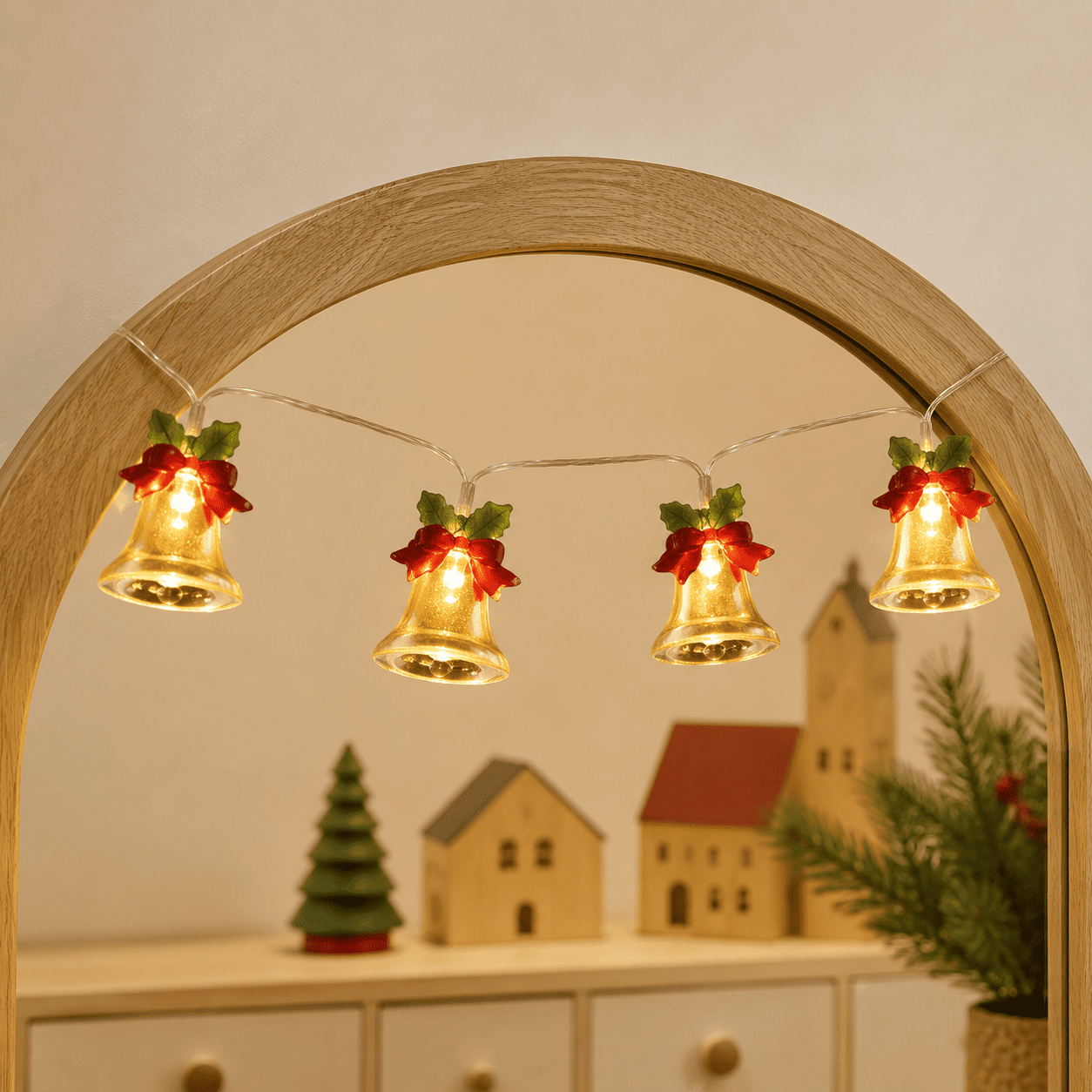 Guirlande lumineuse de Noël à cloches dorées et nœuds rouges, suspendue à un miroir en bois clair.