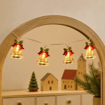 Guirlande lumineuse de Noël à cloches dorées et nœuds rouges, suspendue à un miroir en bois clair.